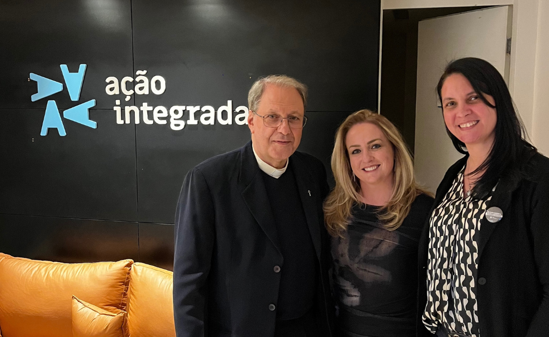 Fotografia de três pessoas em frente ao painel com a identidade visual do Projeto Ação Integrada. Ao lado esquerdo, Monsenhor Manuel Manangão, Diretor Presidente da Cáritas RJ. Ao seu lado, a criadora do Projeto Ação Integrada RJ, Dra. Guadalupe Turos Couto; e ao lado direito, Débora Alves, coordenadora da Cáritas RJ.